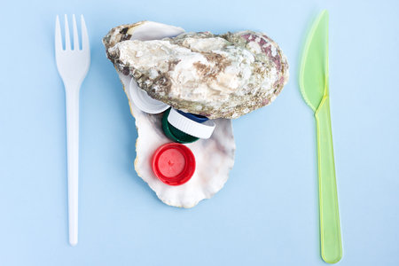 Seashell from oyster with colorful plastic bottle caps, folk and knife. Plastic pollution of food...
