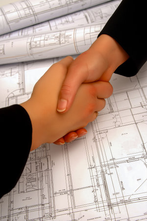 Hands of two individuals shaking over detailed architectural plans on a table, representing collaboration and partnership in construction and design projectsの素材