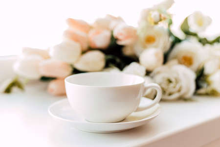 A white cup and saucer stand near the flowers on a light background. High quality photoの写真素材