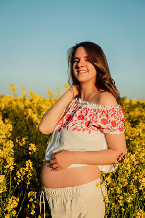 A pregnant woman in a Ukrainian embroidered shirt stands in a rapeseed field