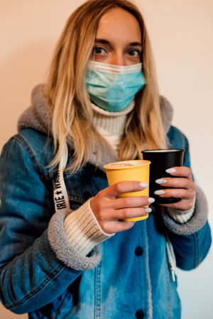 A young woman in a medical mask holds two glasses of coffee in her hands