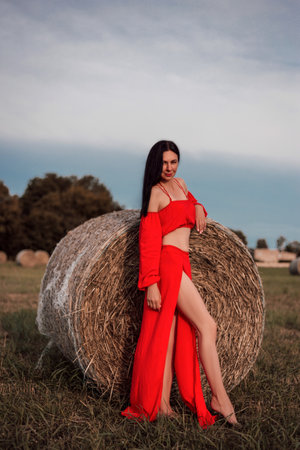 A woman in a red dress stands barefoot in a field near a stack of strawの写真素材