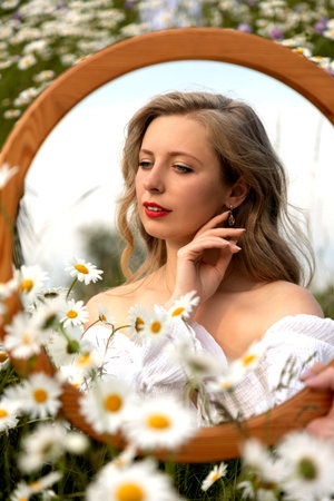 Chamomile field. Woman with a mirror. Photo of young girl face with care of her face in mirrorの写真素材