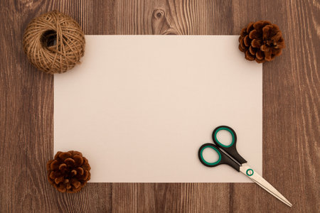 Warm rustic flat lay with blank paper, scissors, pine cones, and twine on a wooden table. Perfect for DIY projects, holiday crafts, or natural handmade designsの写真素材