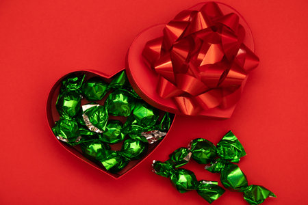 Heart-shaped red box with glossy red bow and green wrapped candies on red background. Bright festive image symbolizing love and celebration.の写真素材