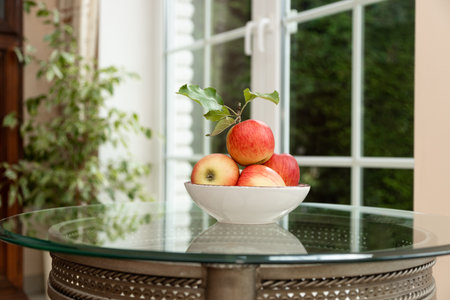 Bowl of ripe red apples on a glass table near a window. Minimalist still life symbolizing healthy eating, apple detox diet, and natural source of vitamins.の写真素材