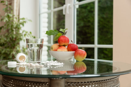 Fresh red apples in a bowl next to pills and a glass of water on a glass table. Symbol of the balance between natural nutrition and medical supplements. Concept of health care, immunity, and wellbeingの写真素材
