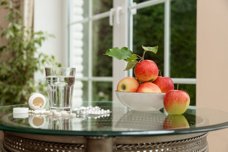 Still life with fresh apples, white pills, and a glass of water on a glass table by the window. Concept of health balance, natural vitamins from fruits versus synthetic supplements, and daily wellness choices.の写真素材