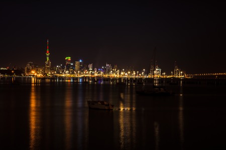 Bright lights of Auckland city downtown and the port reflected in Okahu bay with a small boat in the foreground. North Island, New Zealand. Soft focusの写真素材