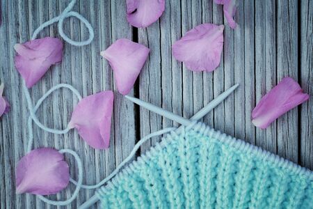 Retro-style photo of knitting needles crossed over knitwork in progress and pink camellia blossom petals on wooden background. Selective focus. Toned imageの写真素材
