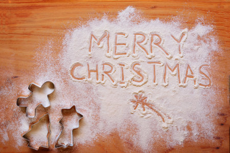 Words 'Merry Christmas' written in spread flour and a variety of cookie cutters on a wooden chopboard. Concept of Holiday Season and festive baking