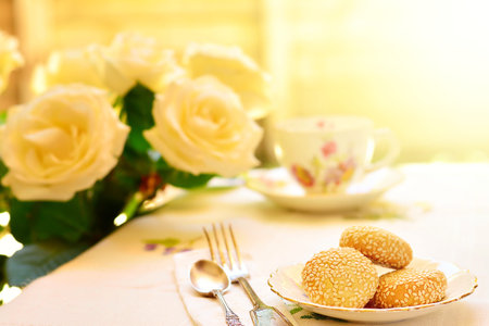 Table set with vintage bone china tea set, sesame biscuits on saucer and white roses on a bright...