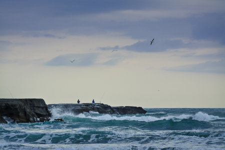 Sunset at Muriwai beach. Stormy waves splashing against the rocks where fishermen bracing the elements and seaguls circling above. Auckland, New Zealandの写真素材