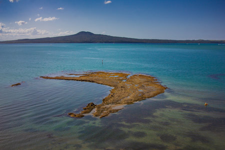 A vibrant view from St Heliers, Auckland, showcasing a rocky tidal flat reaching into the turquoise sea, with Rangitoto Island in the distance under a clear blue sky and scattered clouds.の写真素材