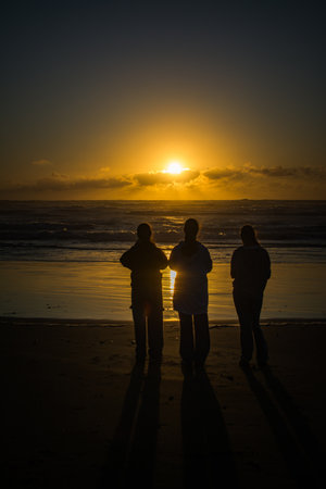 Three young women stand side by side on Muriwai Beach, silhouetted against a glowing sunset, casting long shadows on the sand as the waves roll in under a golden skyの写真素材