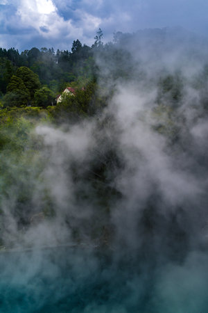 Thick geothermal steam rises from a vivid turquoise pool, partially veiling the surrounding lush forest and a quaint red-roofed house in the distance. Rotoruas surreal volcanic beauty.の写真素材