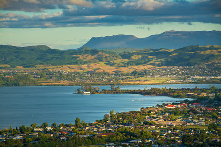 A breathtaking view of Rotorua city nestled beside Lake Rotorua, with rolling green hills and distant mountains under a dramatic sky, showcasing the harmony of nature and communityの写真素材