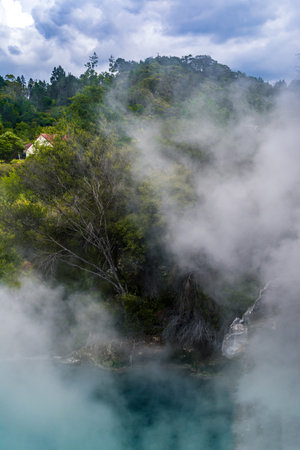 Thick geothermal steam rises from a vivid turquoise pool, partially veiling the surrounding lush forest and a quaint red-roofed house in the distance. Rotorua's surreal volcanic beautyの写真素材