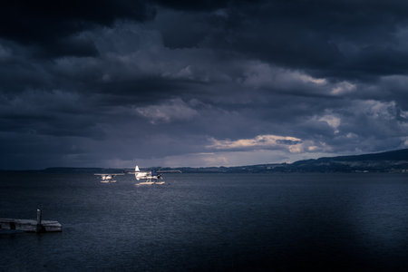 Dramatic storm clouds open to reveal soft pink light over Lake Rotorua, where two seaplanes rest on calm waters, creating a striking contrast of nature power and human adventure.の写真素材