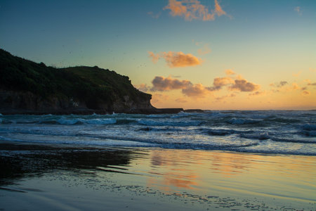 Waves roll gently onto the reflective sands of Muriwai Beach at dusk, as the last light of sunset paints the clouds and sea with soft hues of orange, pink, and blueの写真素材