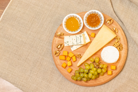Assorted cheese on a round wooden plate Camembert cheese, slices of hard cheese, walnuts, grapes, honey, apricot jamの写真素材