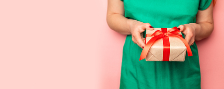 Holiday greetings concept. Female hands hold surprise crafting box with red ribbon bow on a pink background with place for textの写真素材