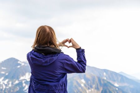 Young woman making heart shape with hands looks at the nature green hills of the mountains and enjoys the view. Amazing nature conceptの写真素材
