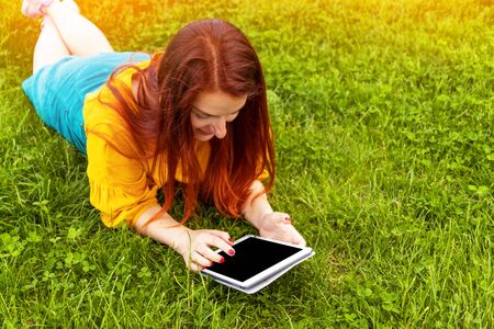 Happy young red-haired girl in a yellow jacket and green skirt lying on the grass and using a tablet, watching an educational video in a park outdoors.の写真素材