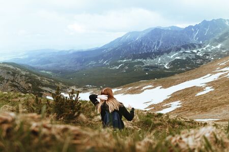 Happy young blonde travels with a blue backpack, sits on top of a mountain and enjoys green mountain scenery. Back viewの写真素材