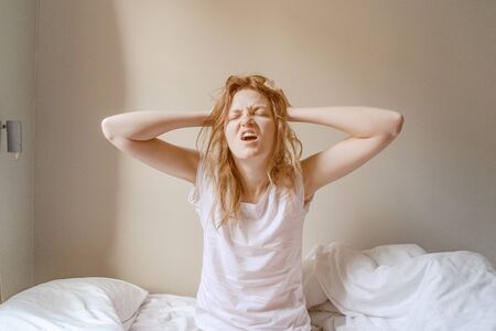 Bad Morning. Frowning blonde in bed with hands on face. Young woman having trouble with getting up early in the morningの写真素材