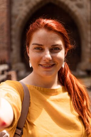 Red-haired young girl in a yellow sweater takes a selfie against the background of the sights on the street on a summer day.の写真素材