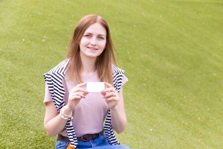 Young smiling woman holding a blank business card sitting on a wooden bench in the park on a summer dayの写真素材