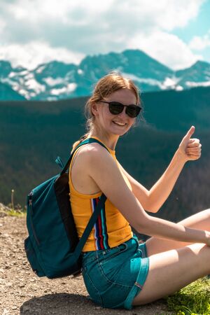 Blonde cute girl with a backpack and glasses sits on a mountain, shows a sign the class is enjoying the beautiful hills of the mountains on a sunny day.の写真素材