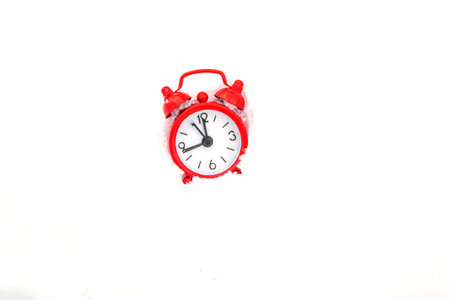 Red metal alarm clock clock in the snow. The hand on the clock shows 12 oclock
