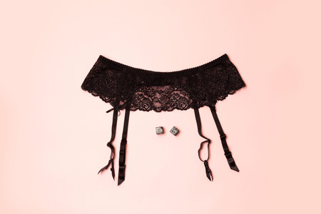 Top view of black lace belt on hips, playing cube on a pink background. Adult game concept