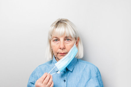 Adult blond woman in a blue shirt with a medical mask against a gray wall. Protection against...
