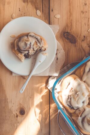 Freshly baked cinnamon buns with cream in a glass dish on a wooden table, top view flat layの写真素材