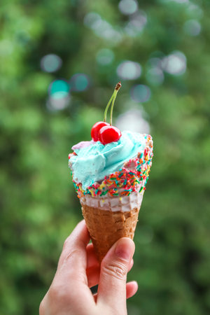 Female hand holds multi-colored ice cream with cherries in a waffle cone against blur green...