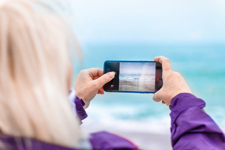 Tourist caucasian blond woman traveler taking a picture of sunset by phone on the beach by the...
