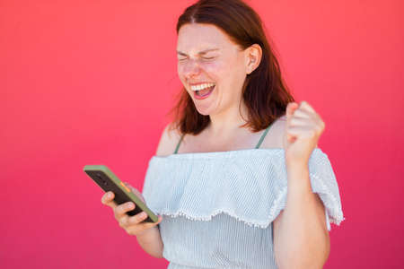 Happy young girl looking at smartphone screen and winning internet on pink wall background, celebrating a victoryの写真素材