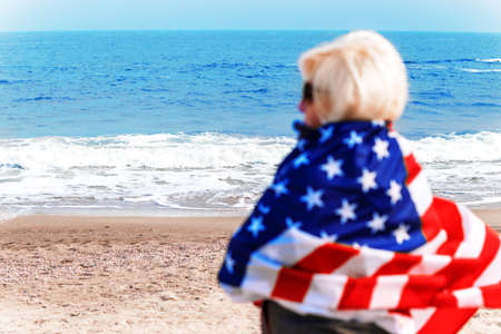 Caucasian blonde woman standing completely wrapped in american flag and looking at sea. Usa flag for independence dayの写真素材