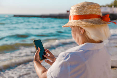 Portrait beautiful caucasian blond woman in straw hat and striped shirt makes photo by smartphone of the sea on a sunny day.の写真素材
