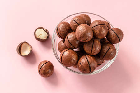 Raw macadamia nuts in a transparent glass bowl and scattered on pink solid background. Minimal composition.の写真素材