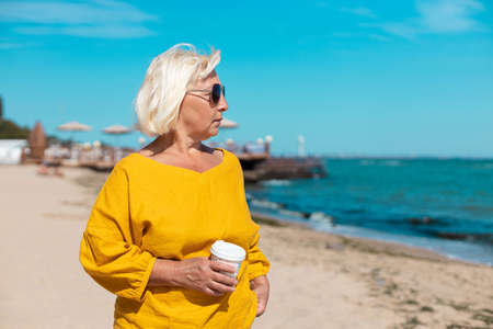 Beautiful woman holding paper cup of coffee and enjoying a walk along the sea coastの写真素材
