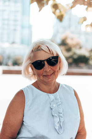 Portrait of adult blonde positive satisfied mature woman in sunglasses smiling at camera outdoors in...