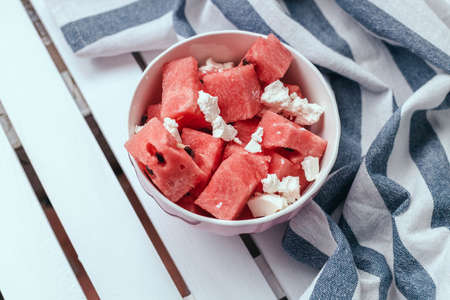 Fresh summer salad with slices of watermelon and feta cheese on a white bowl with a striped towel on a wooden white backgroundの写真素材