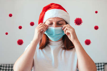 Young woman in Santa Claus hat puts on a protective medical mask while staying at home. Christmas covid 19 conceptの写真素材