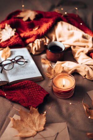 Burning candles, cup of hot tea, open book and glasses on plaid in bed. Autumn leaves and light garland decor on sofa. Autumn style. Hygge concept, vertical photo orientationの写真素材