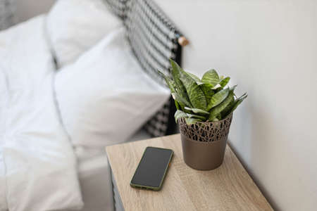 Black smartphone on the nightstand. Green plant in a flower pot on the table in the bedroom against the background of the bedの写真素材