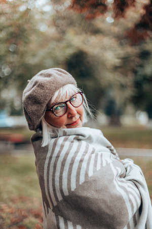 Happy caucasian lady in stylish beret,glasses, cozy blanket walk on a windy day in autumn or fallの写真素材